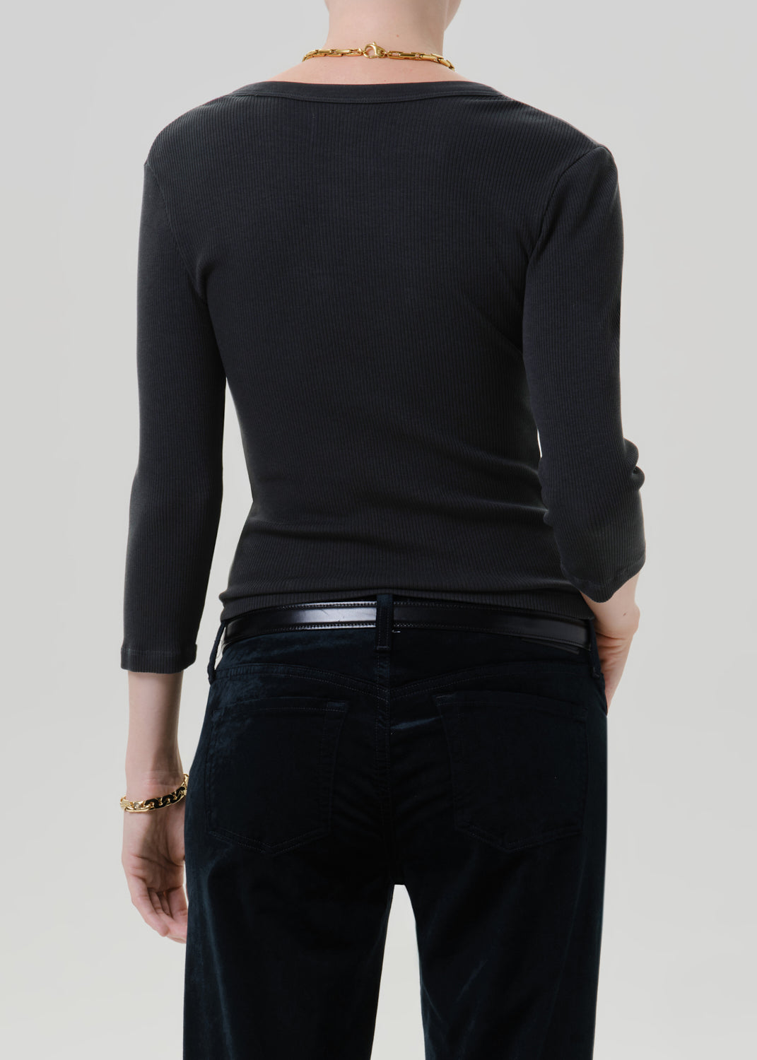 Person wearing a black long sleeve fitted henley top with black jeans, back cropped view on a grey background.