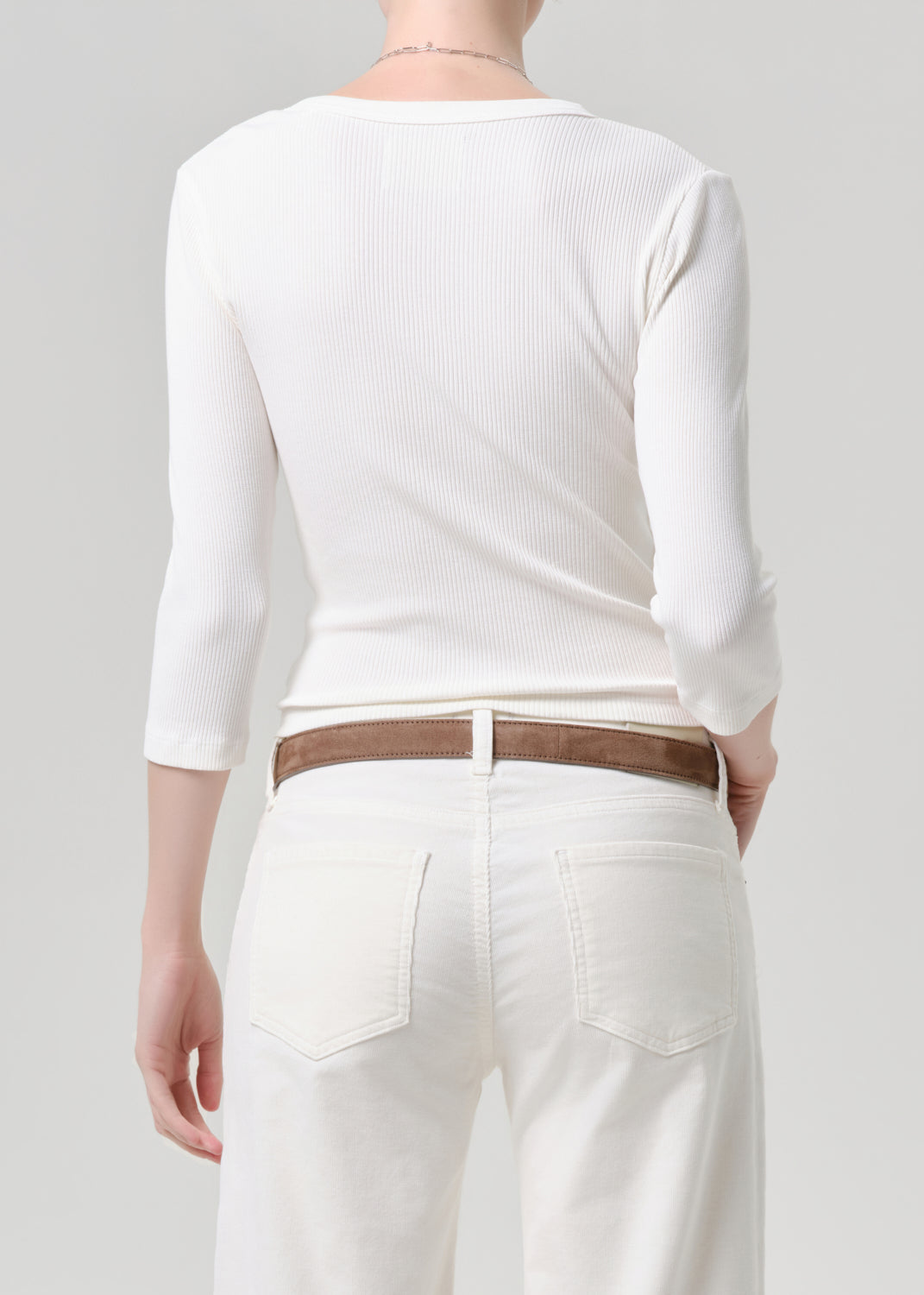 Person wearing a white long-sleeve top and white pants with a brown belt on a grey background
