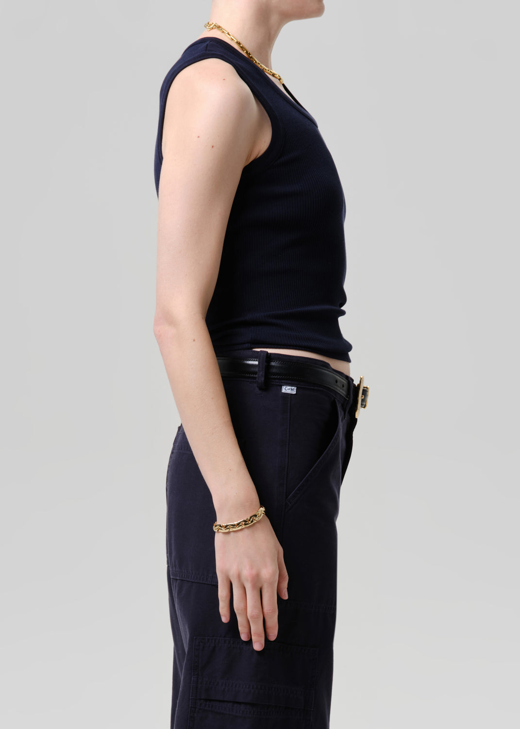 Person wearing a black sleeveless top and pants with gold jewelry on a grey background