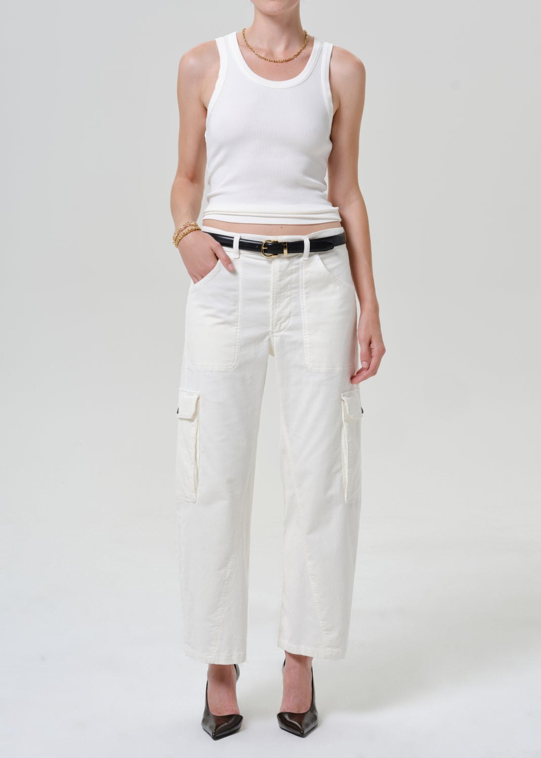 A model wearing a white ribbed tank top, white cropped cargo pants with a black belt, and black heels, standing against a light gray studio background.