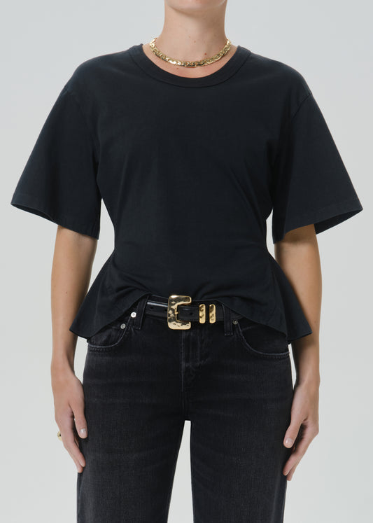 A model wearing a black short-sleeve crewneck t-shirt tucked into black jeans with a gold belt, against a grey background.