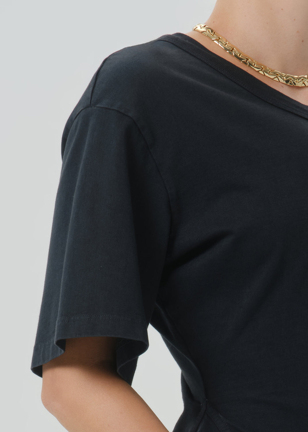 Close-up of a person wearing a black short-sleeve crew-neck T-shirt and a chunky gold chain necklace against a grey background.