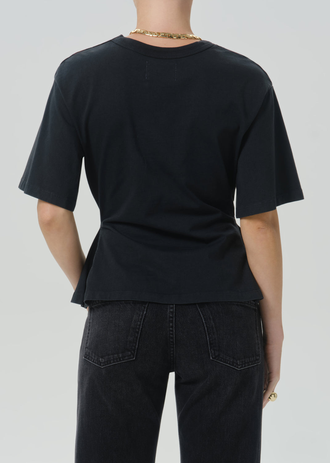 Rear view of a person wearing a black short-sleeve crew-neck T-shirt and dark jeans against a grey background.