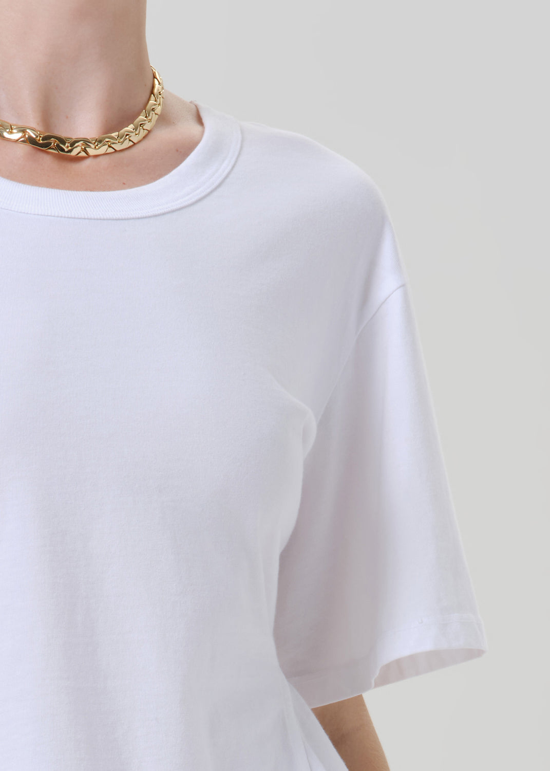 Close-up of a person wearing an oversized white short-sleeve crewneck t-shirt and a chunky gold chain necklace, set against a solid grey background.