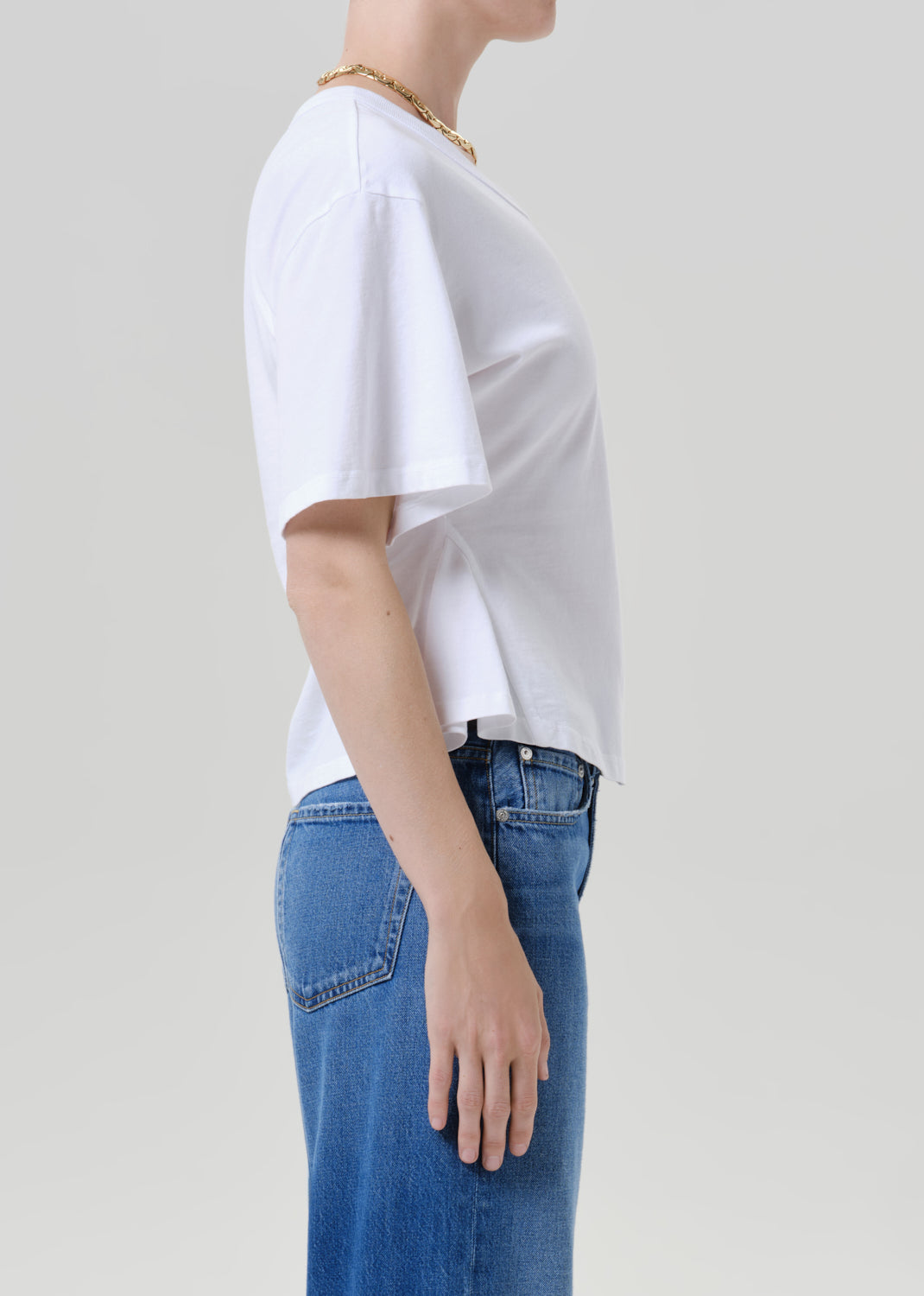 A side-profile view of a person wearing a white short-sleeve crop top and blue jeans against a grey background.