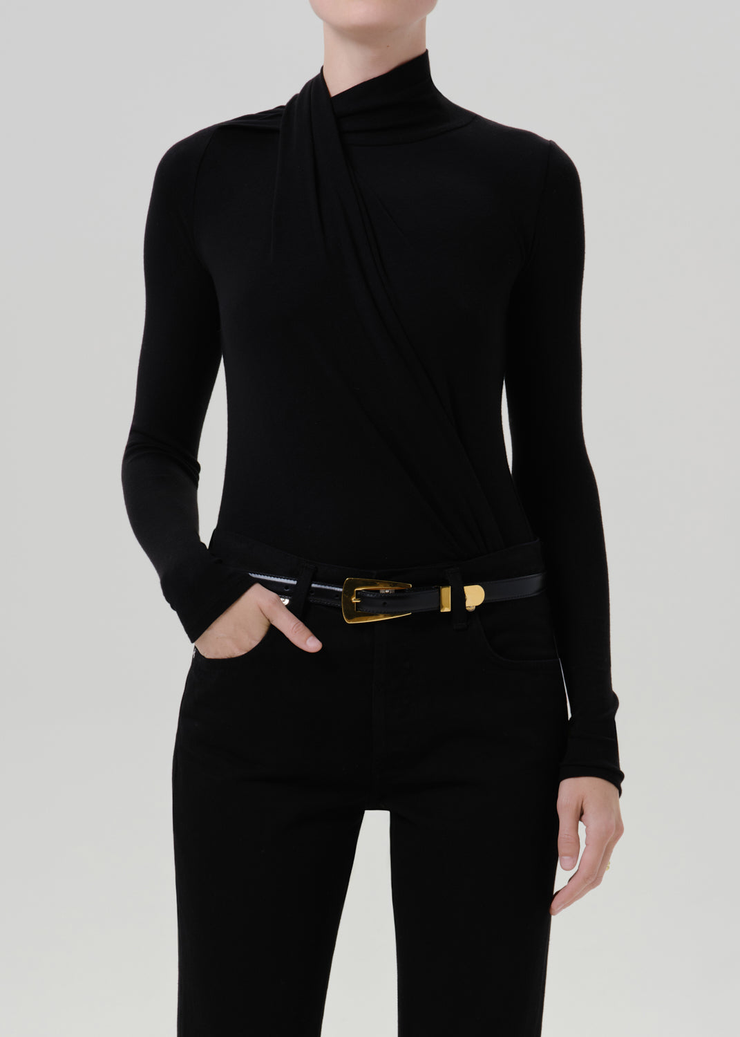 Person wearing a black long sleeve draped mock neck top with black pants on a grey background