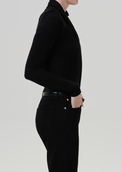 Person wearing a black long sleeve draped mock neck top with black pants viewed from the side on a grey background