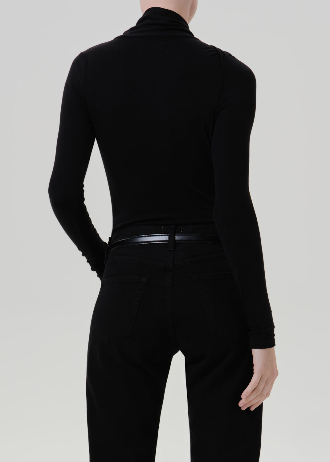 Person wearing a black long sleeve draped mock neck top with black pants viewed from the back on a grey background