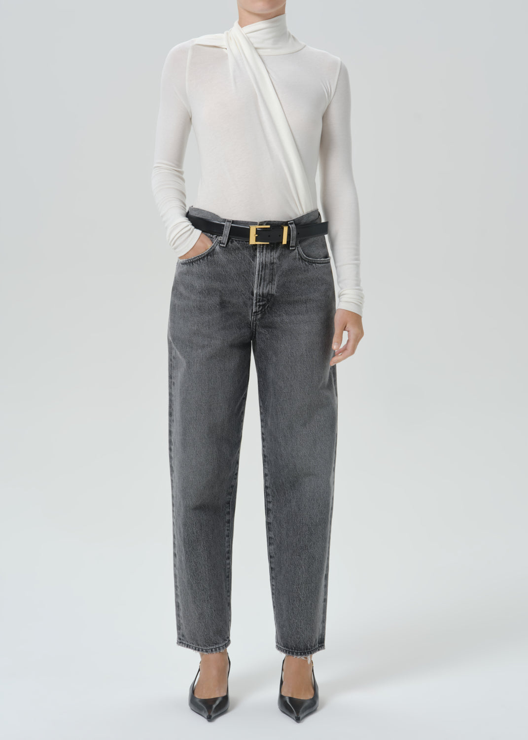 A model wearing a white asymmetrical long-sleeve top, dark gray wide-leg jeans with a black belt featuring a gold buckle, and black pointed-toe heels, photographed against a grey background.