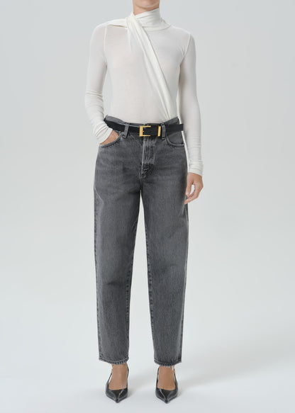 A model wearing a white asymmetrical long-sleeve top, dark gray wide-leg jeans with a black belt featuring a gold buckle, and black pointed-toe heels, photographed against a grey background.