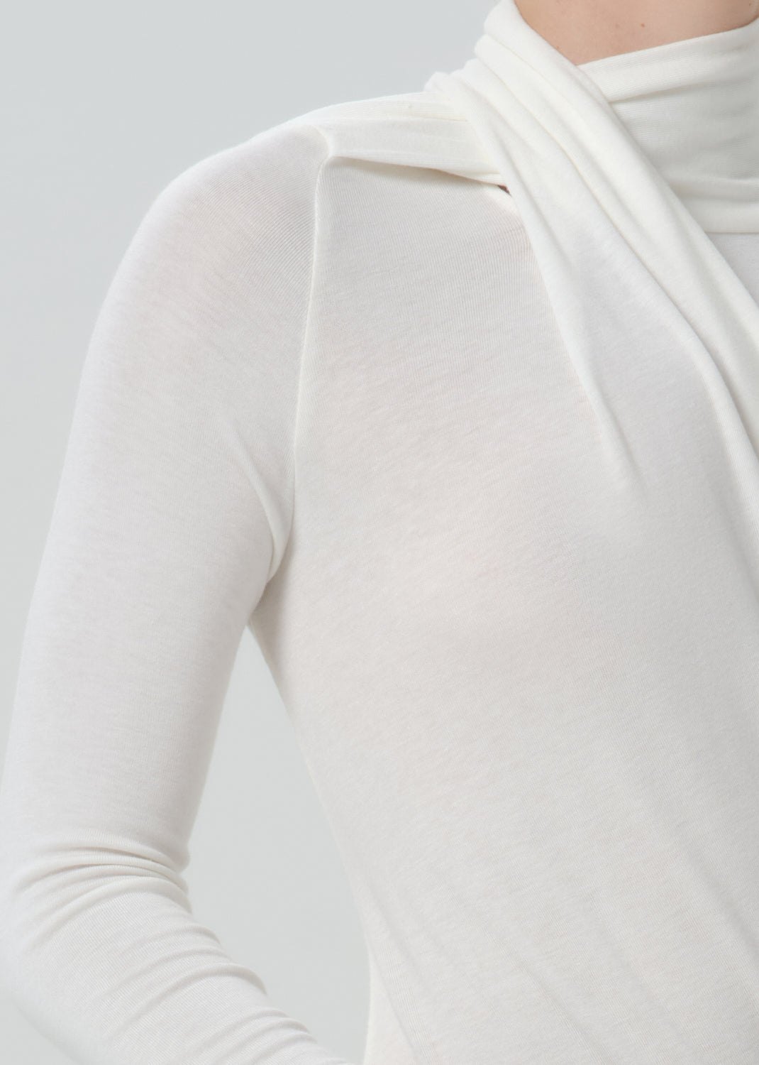 Close-up of a person wearing a fitted long-sleeve top with a draped mock neck in a warm off-white or cream color, against a grey background.