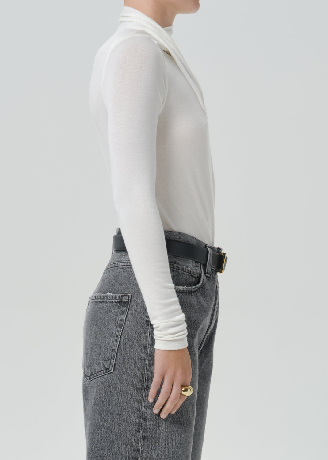 A side profile view of a model wearing a white long-sleeve mock neck top tucked into high-waisted grey jeans with a black belt.