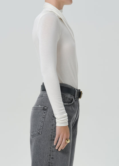 A side profile view of a model wearing a white long-sleeve mock neck top tucked into high-waisted grey jeans with a black belt.