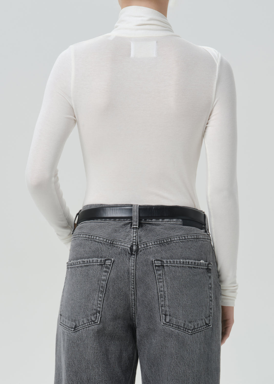 Back view of a model wearing a white long-sleeve mock neck turtleneck top and gray washed jeans with a black belt, against a grey background.