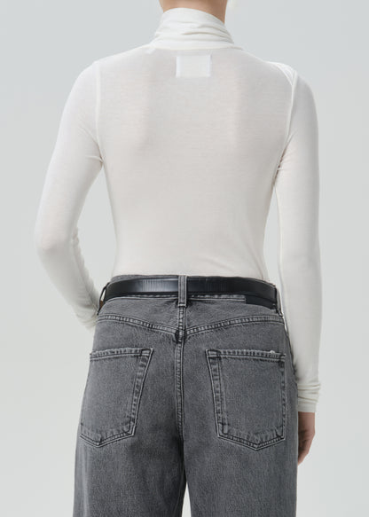 Back view of a model wearing a white long-sleeve mock neck turtleneck top and gray washed jeans with a black belt, against a grey background.