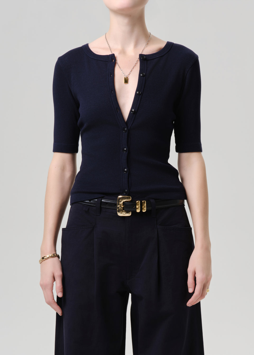 Person wearing a black top and black pants with a gold belt on a grey background