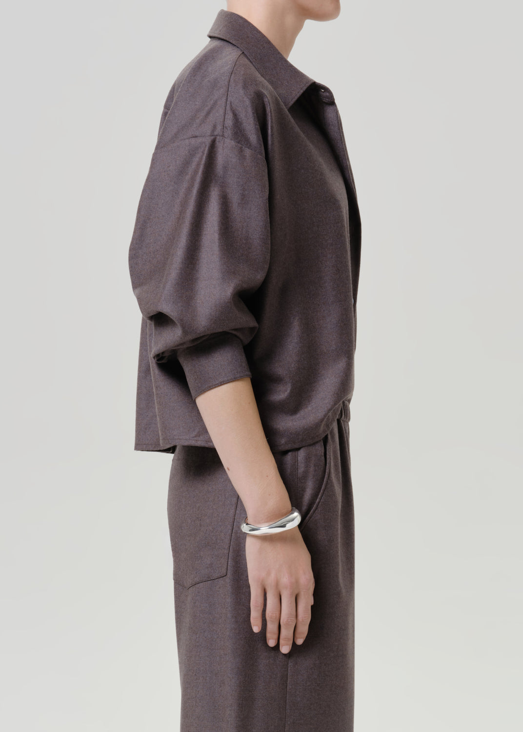 Woman wearing a dark brown short sleeve button-up shirt with matching dark brown wide leg pants viewed from the side on a grey background.