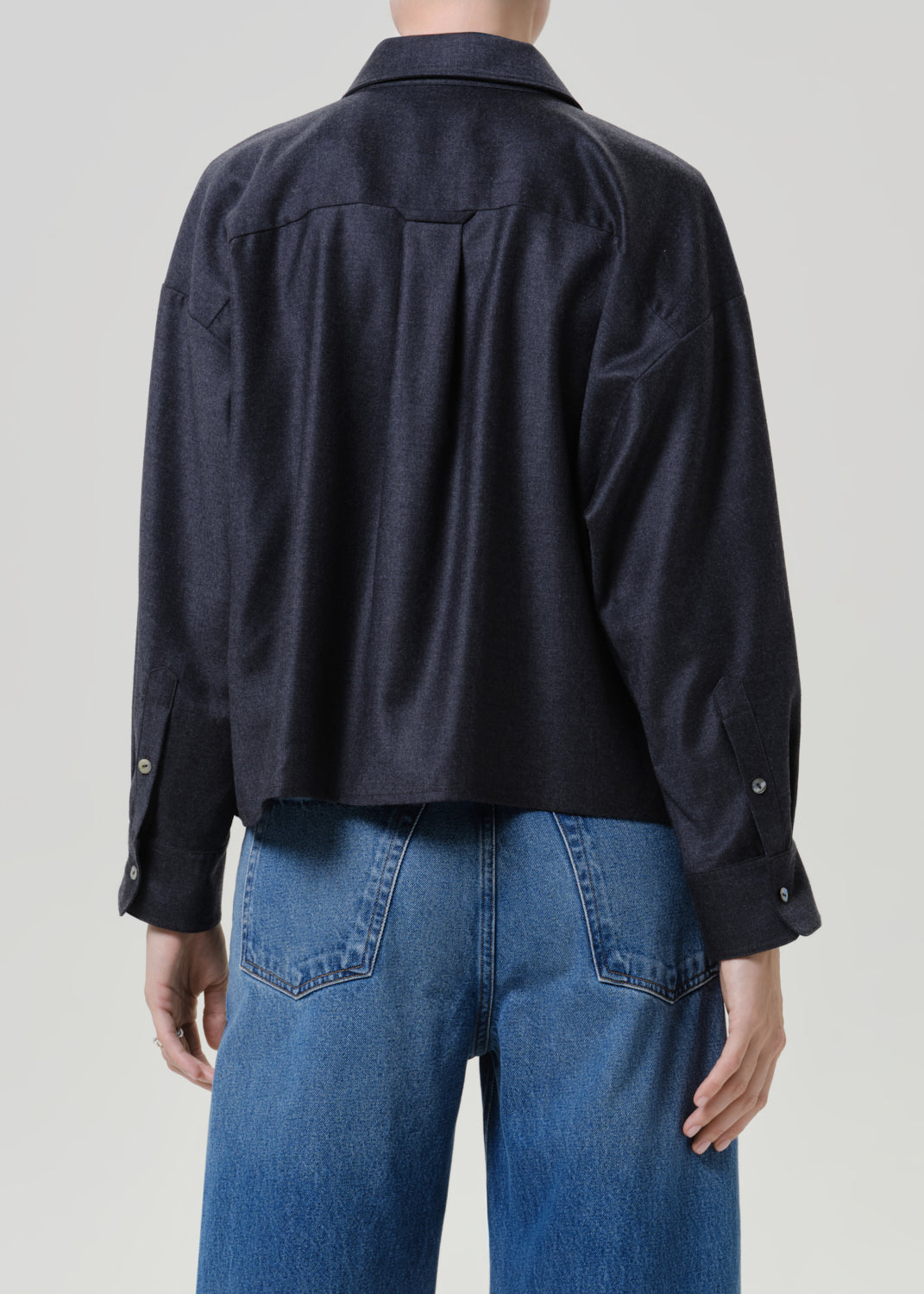Woman wearing a dark navy button-up shirt with medium blue jeans viewed from the back on a grey background.
