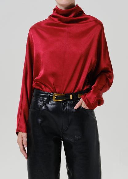 Woman wearing a red satin draped neck top with black leather pants on a grey background.