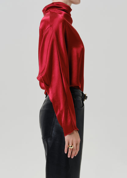 Woman wearing a red satin draped neck top with black leather pants viewed from the side on a grey background.