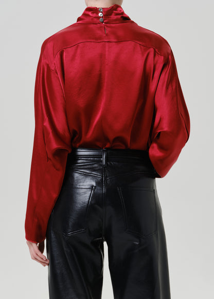 Woman wearing a red satin draped neck top with black leather pants viewed from the back on a grey background.