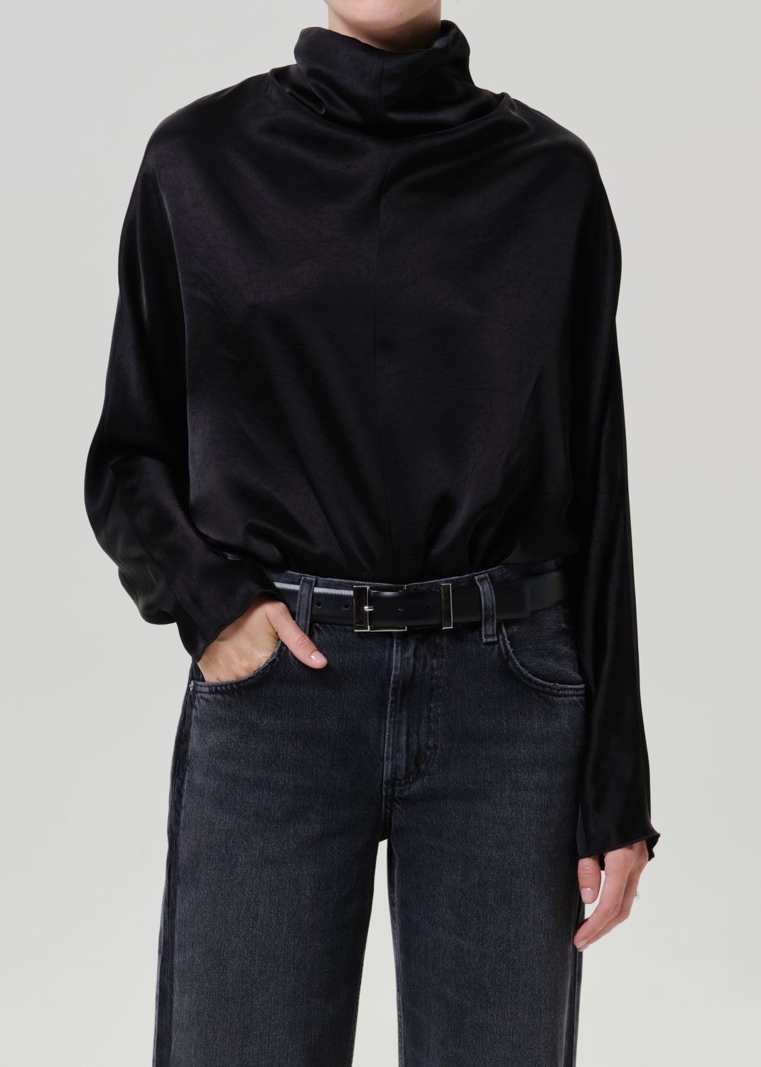 Woman wearing a black satin draped neck top with black jeans on a grey background.