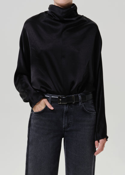 Woman wearing a black satin draped neck top with black jeans on a grey background.