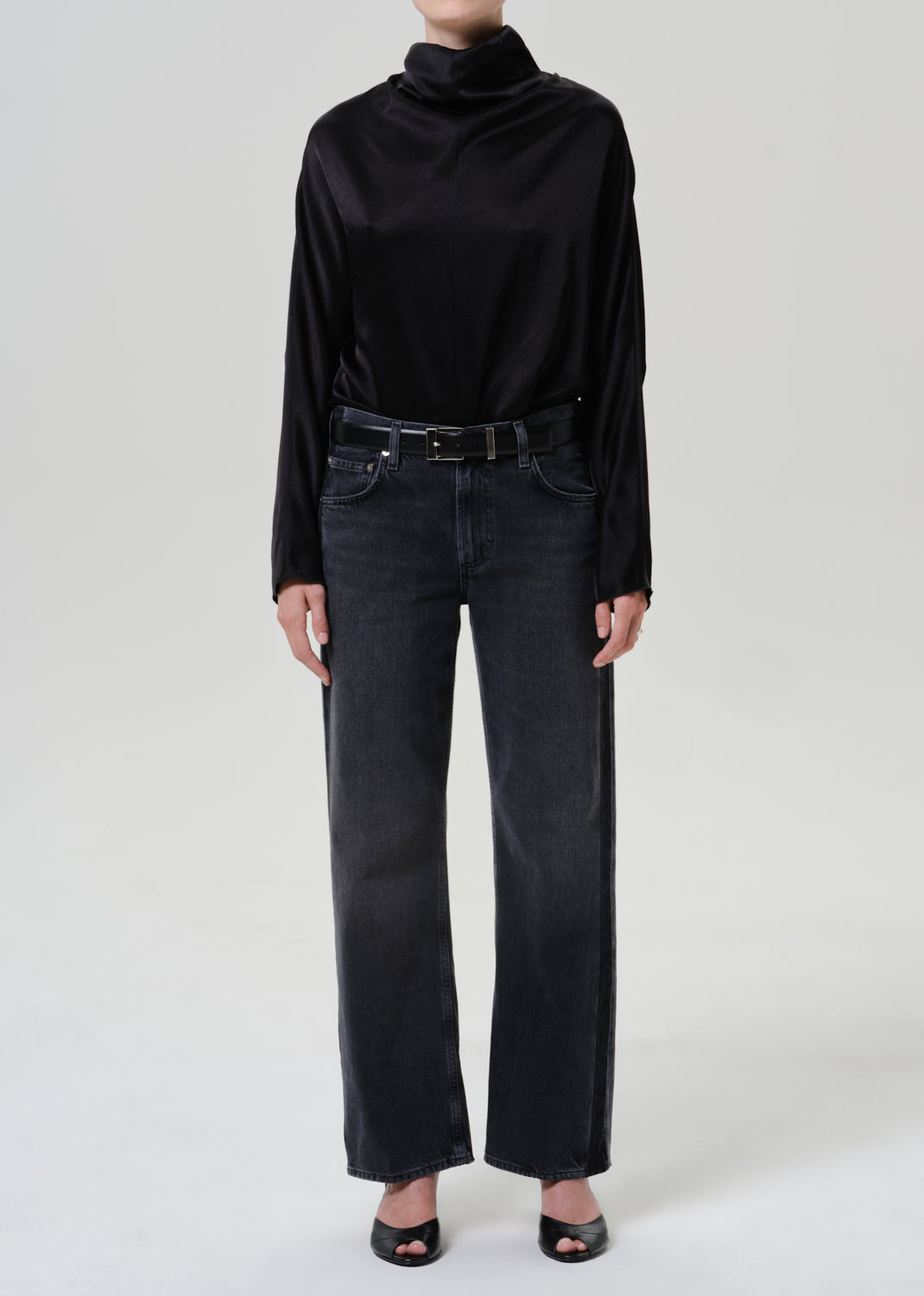 Woman wearing a black satin draped neck top with black jeans on a grey background.