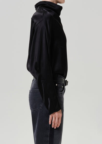 Woman wearing a black satin draped neck top with black jeans viewed from the side on a grey background.