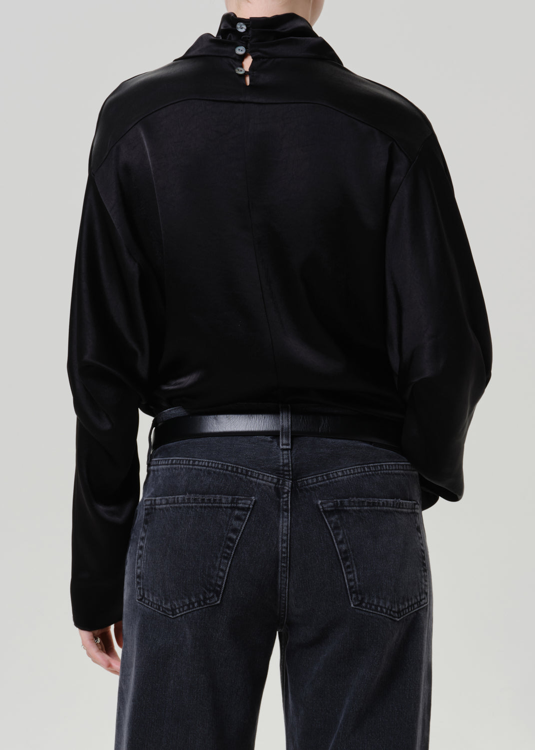 Woman wearing a black satin draped neck top with black jeans viewed from the back on a grey background.