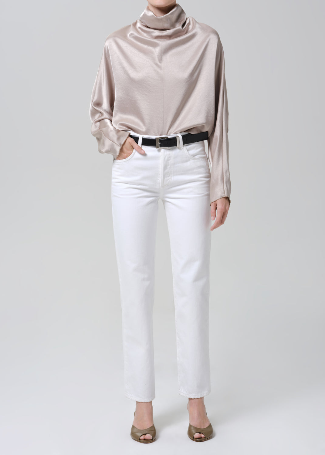 Woman wearing a taupe satin draped neck top with white straight leg pants on a grey background.