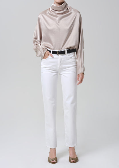 Woman wearing a taupe satin draped neck top with white straight leg pants on a grey background.