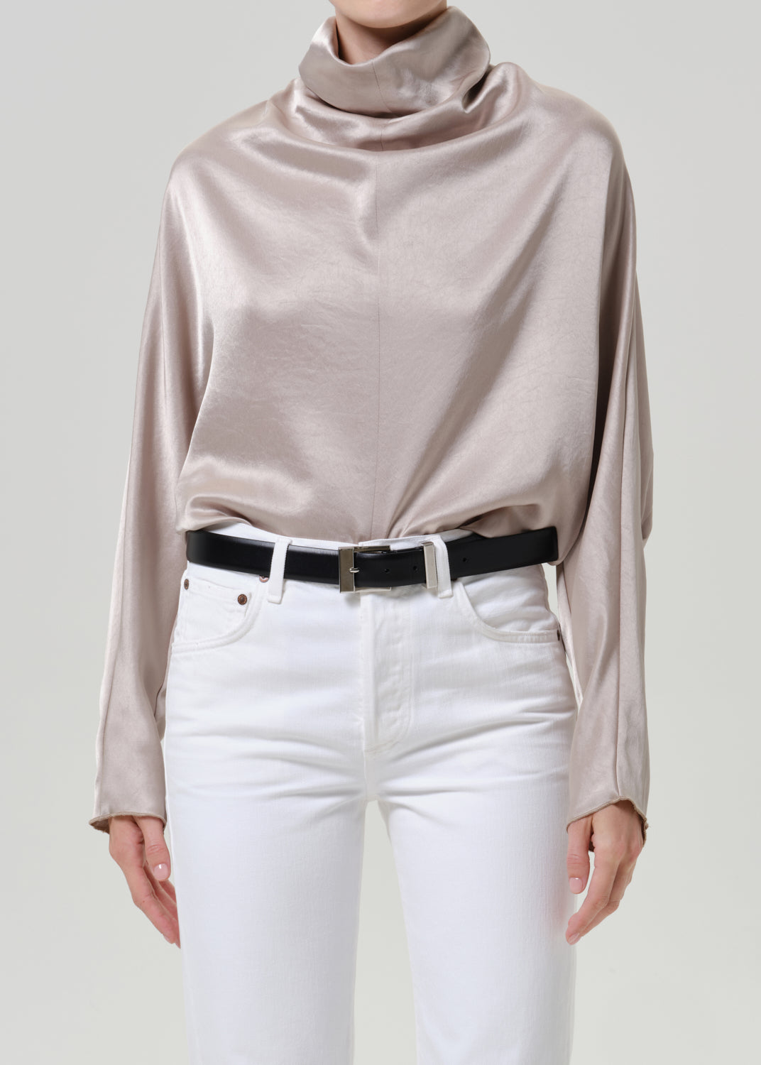 Woman wearing a taupe satin draped neck top with white straight leg pants on a grey background.