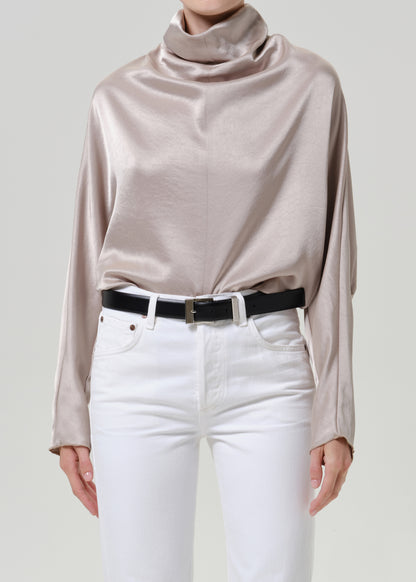 Woman wearing a taupe satin draped neck top with white straight leg pants on a grey background.