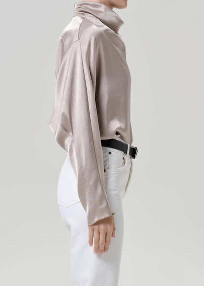 Woman wearing a taupe satin draped neck top with white straight leg pants viewed from the side on a grey background.
