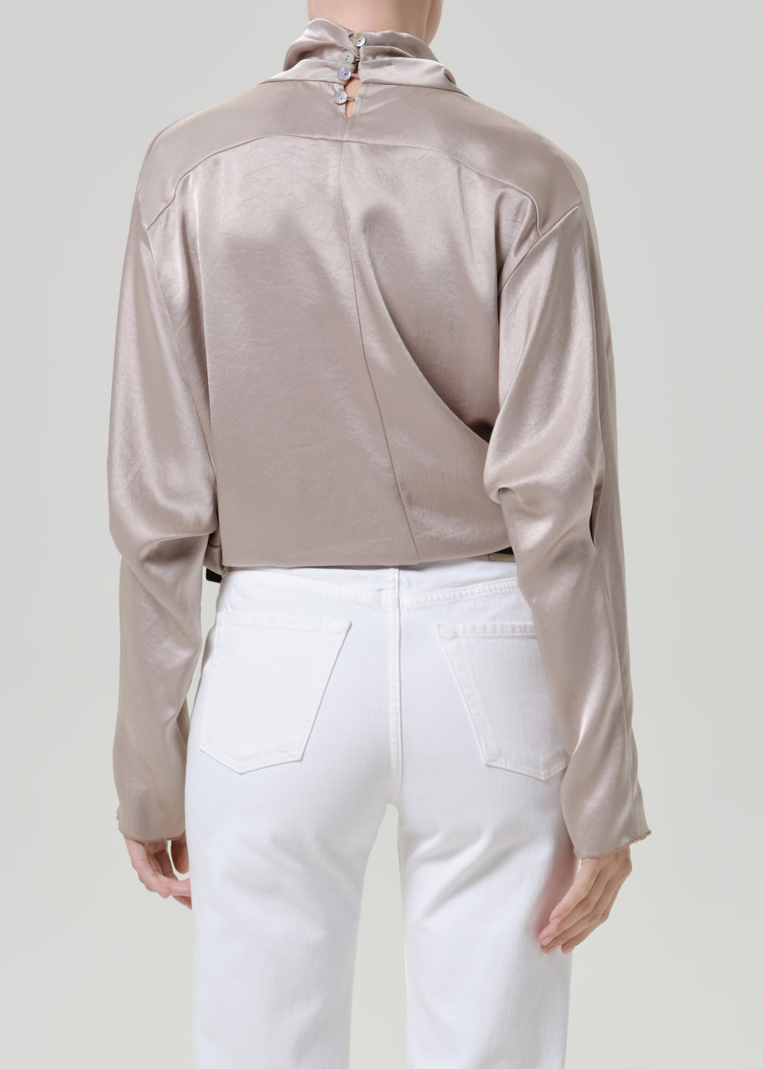 Woman wearing a taupe satin draped neck top with white straight leg pants viewed from the back on a grey background.