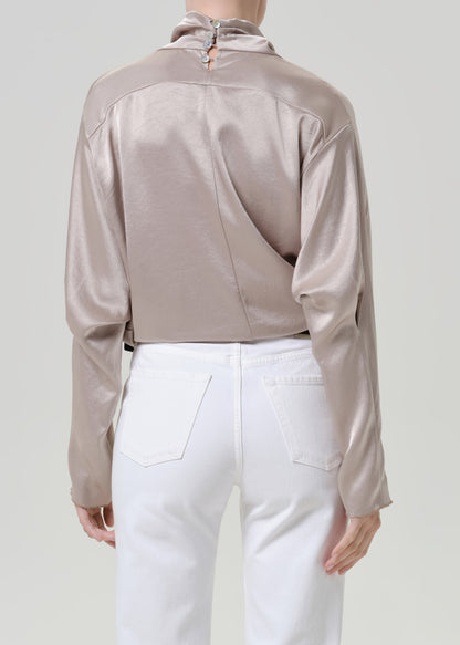 Woman wearing a taupe satin draped neck top with white straight leg pants viewed from the back on a grey background.