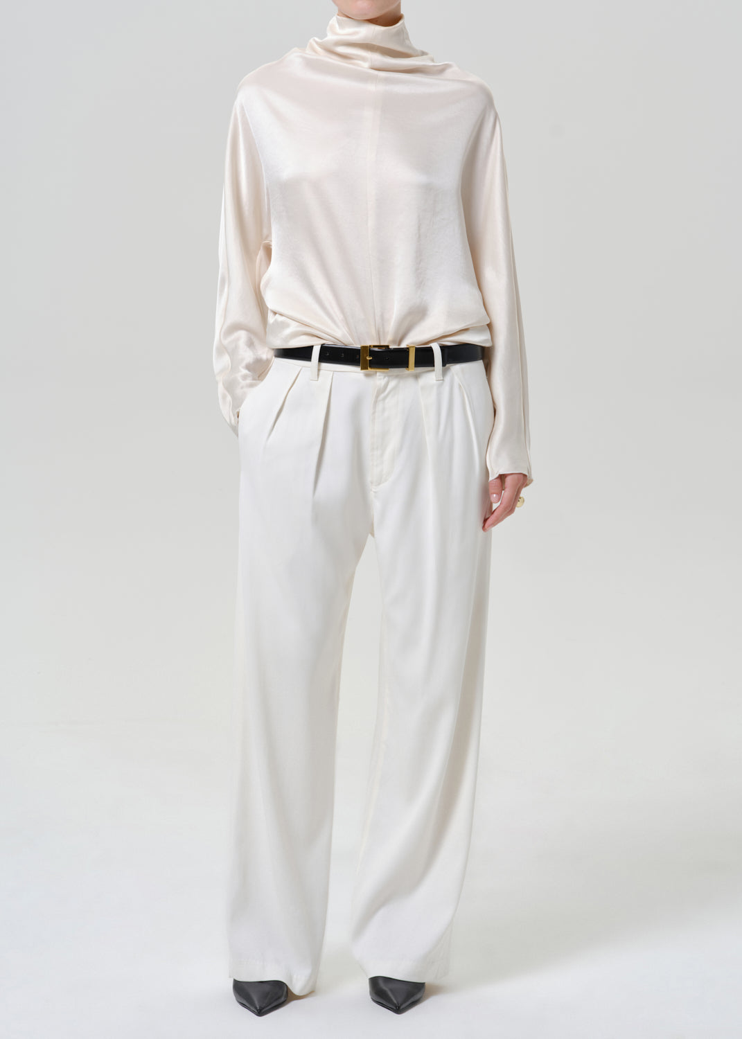 Woman wearing an ivory satin draped neck top with white wide leg pants on a grey background.