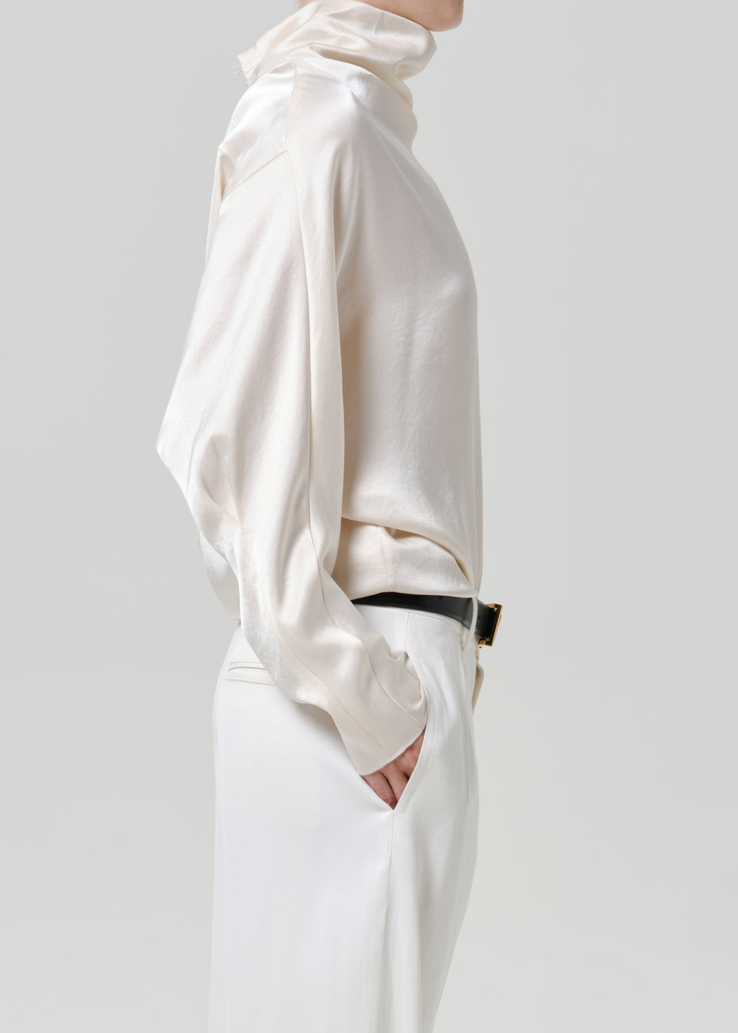 Woman wearing an ivory satin draped neck top with white wide leg pants viewed from the side on a grey background.