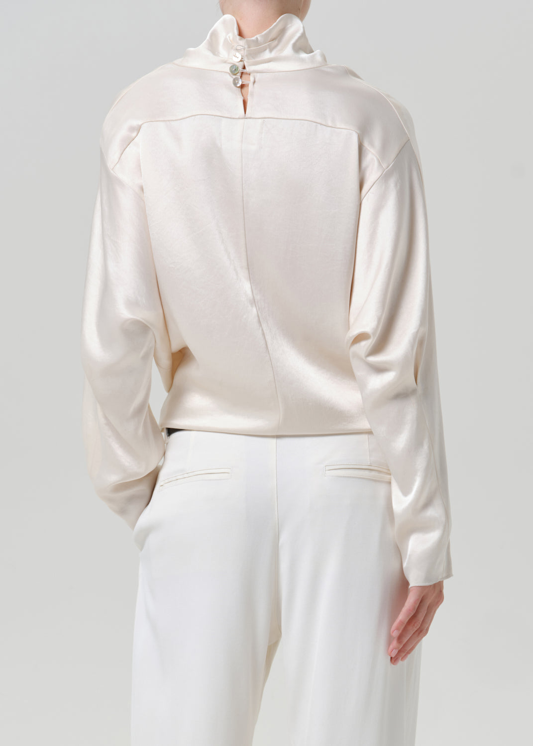 Woman wearing an ivory satin draped neck top with white wide leg pants viewed from the back on a grey background.