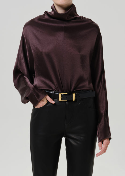 Woman wearing a dark brown satin draped neck top with black leather pants on a grey background.