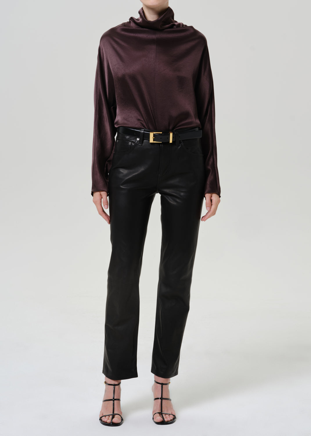Woman wearing a dark brown satin draped neck top with black leather pants on a grey background.