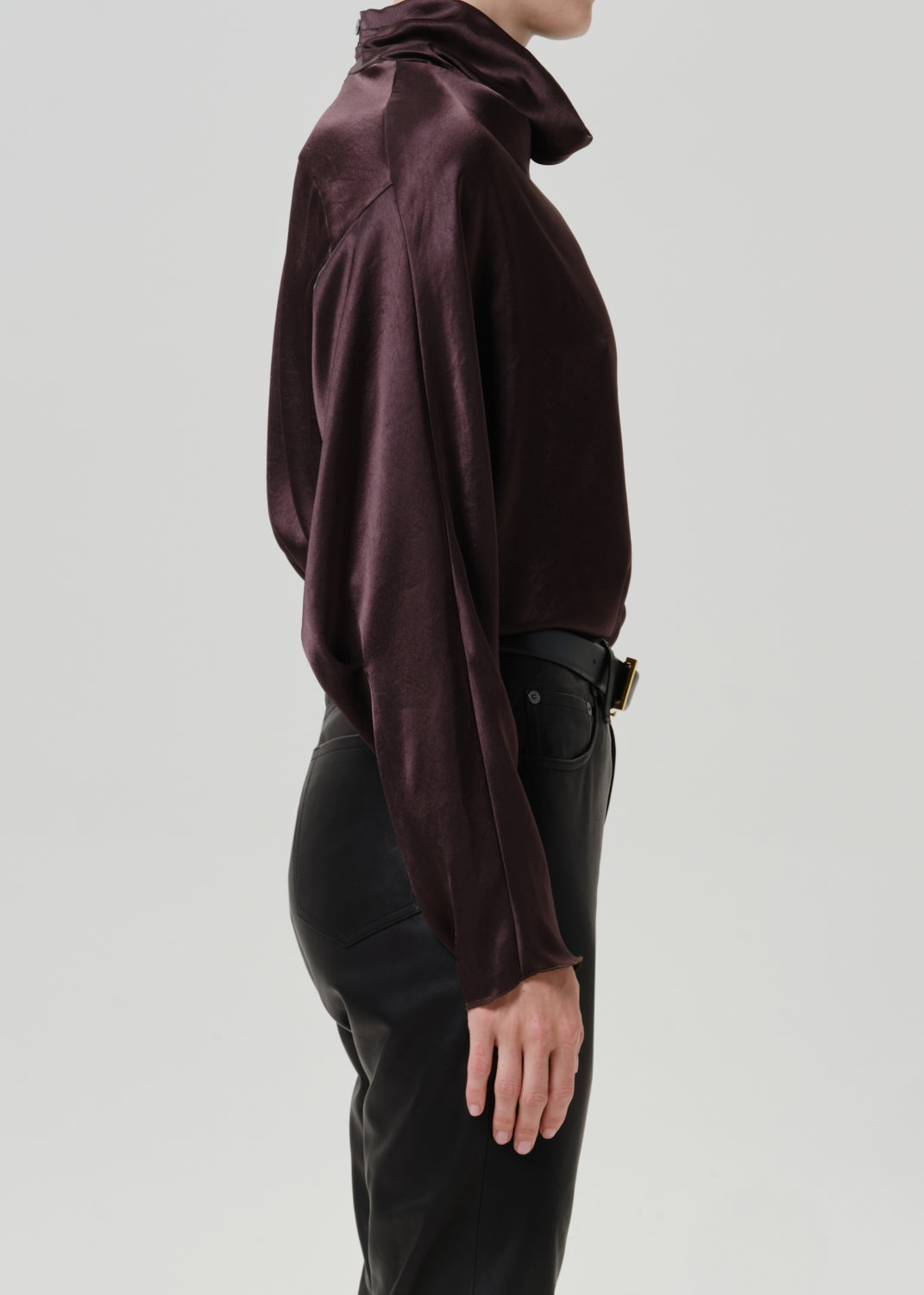 Woman wearing a dark brown satin draped neck top with black leather pants viewed from the side on a grey background.