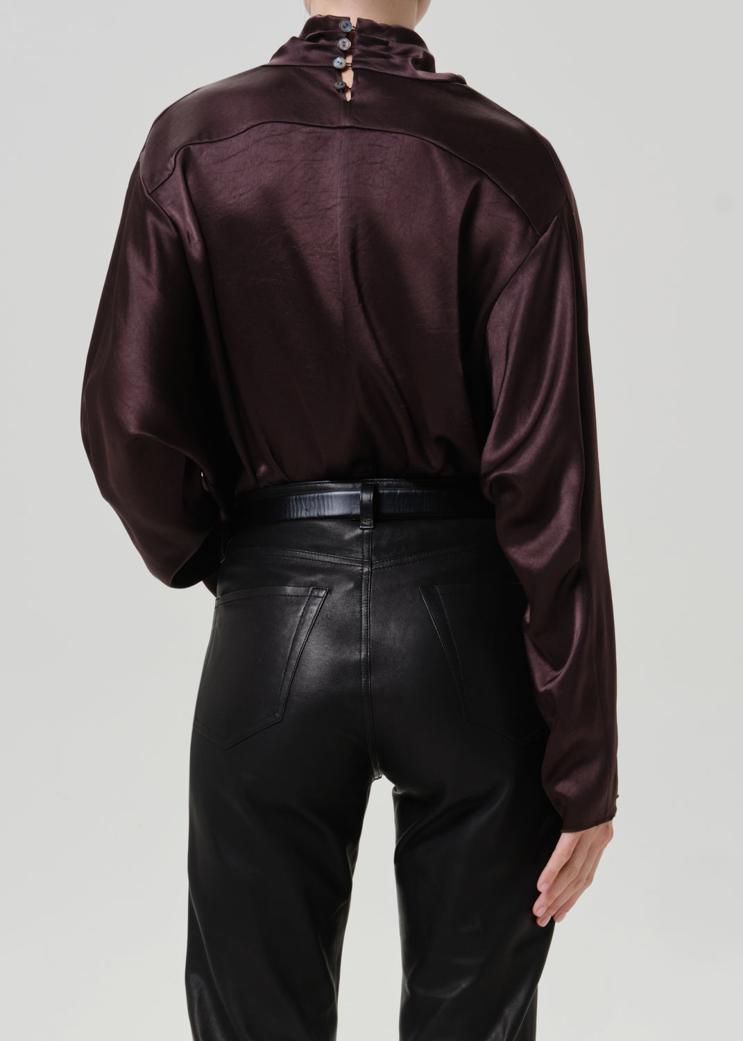 Woman wearing a dark brown satin draped neck top with black leather pants viewed from the back on a grey background.