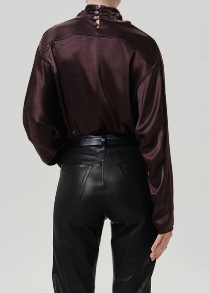 Woman wearing a dark brown satin draped neck top with black leather pants viewed from the back on a grey background.