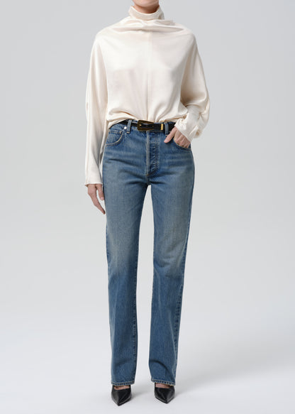 Person wearing an ivory draped cowl neck blouse with blue straight leg jeans on a grey background