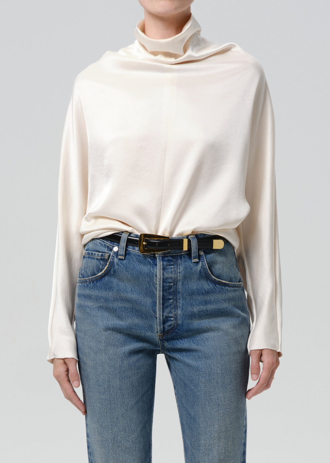 Person wearing an ivory draped cowl neck blouse with blue straight leg jeans on a grey background
