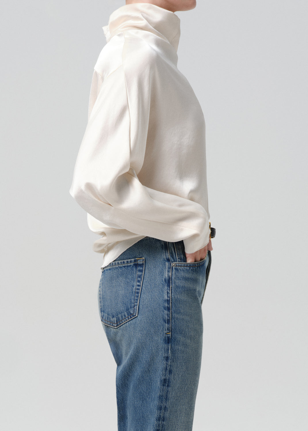 Person wearing an ivory draped cowl neck blouse with blue straight leg jeans viewed from the side on a grey background