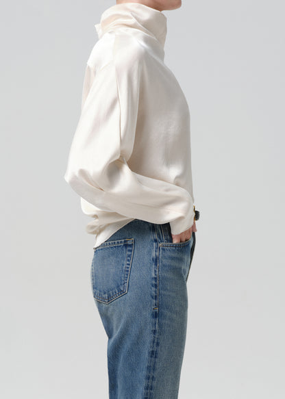 Person wearing an ivory draped cowl neck blouse with blue straight leg jeans viewed from the side on a grey background