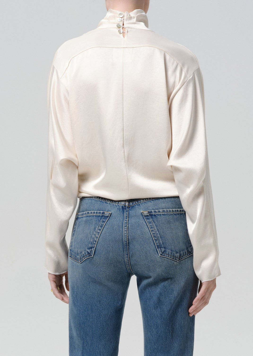Person wearing an ivory draped cowl neck blouse with blue straight leg jeans viewed from the back on a grey background